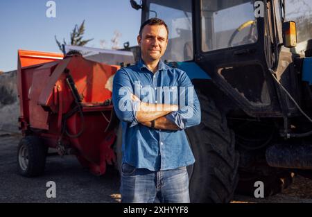 Un giovane con abiti casual si trova accanto ai trattori di un'azienda agricola. Un'attraente macchina agricola e agricola. Un conducente di trattore sorridente dopo il campo Foto Stock