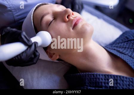 Una giovane donna in accappatoio mente e riceve una procedura di pulizia del viso utilizzando un dispositivo presso un centro benessere o un centro di bellezza. Un volto femminile naturale con una guarigione Foto Stock