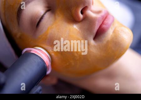 Una giovane donna in accappatoio mente e riceve una procedura di pulizia del viso utilizzando un dispositivo presso un centro benessere o un centro di bellezza. Un volto femminile naturale con una guarigione Foto Stock