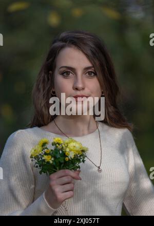 Ritratto di una bella ragazza sorridente con capelli scuri e grandi occhi. Ha in mano un piccolo mazzo di fiori d'autunno gialli. Foto Stock