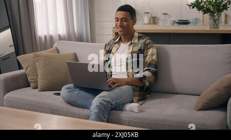 Uomo afro-americano sorridente che si rilassa nell'appartamento sul divano ragazzo maschio etnico birazziale sul divano utente di mutuo ipotecario libero professionista studente uomo d'affari che usa Foto Stock