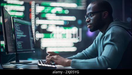 Programmatore maschio nero che lavora nella sala di monitoraggio, circondato da grandi schermi che visualizzano linee di codice del linguaggio di programmazione. Ritratto dell'uomo che crea un software. Concetto futuristico di codifica. Foto Stock