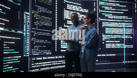 Ritratto di due diversi sviluppatori che utilizzano un notebook, discutendo delle linee di codice che appaiono sui grandi schermi che li circondano. Programmatori maschi e femmine che creano un software insieme, correggono i bug Foto Stock