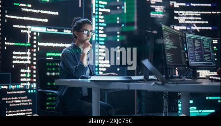 Donna concentrata che lavora su un computer, digita righe di codice che appaiono sui grandi schermi che la circondano in una sala di monitoraggio. Programmatore femminile che crea software innovativo utilizzando dati e sistemi ai Foto Stock