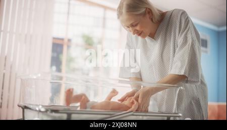 Madre che si lega con il suo bambino neonato vicino ospedale Bassinet letto in una moderna clinica infermieristica. Prendersi cura della mamma tenendo e confortando il suo neonato. Concetto di assistenza sanitaria, gravidanza e maternità Foto Stock