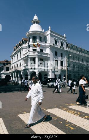 Centro citta' di Kandy, Sri Lanka, lo storico Queens Hotel in stile coloniale bianco sullo sfondo. Un uomo in abiti tradizionali che attraversa la strada. Foto Stock