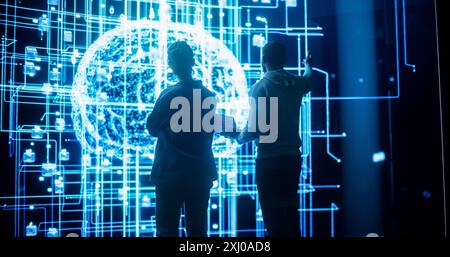 Vista posteriore di due diversi colleghi che guardano un grande schermo con la visualizzazione 3D della rete neurale. Scienziati professionisti dei dati informatici che discutono e lavorano sul monitoraggio delle informazioni private Foto Stock