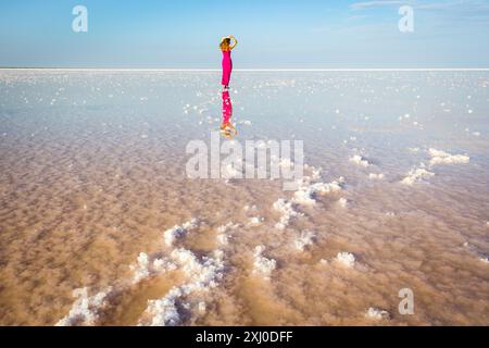 La giovane donna in abito rosso rimane sulla superficie del lago salato con grumi di sale. Foto Stock