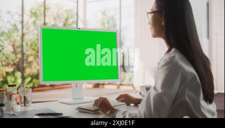 Giovane dottoressa donna che indossa il cappotto bianco, che lavora su un personal computer con il Green Screen Mock Up display nel suo ufficio. Bello asiatico medico sanitario professionista che lavora con i risultati del test Foto Stock