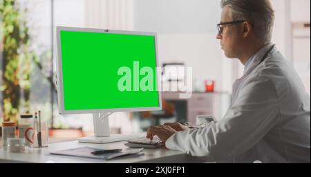 Medico sanitario che lavora sul computer desktop con schermo verde Mock Up display. Professionista della clinica che utilizza PC per efficiente lavoro medico online in ufficio moderno nella struttura di sanità pubblica Foto Stock