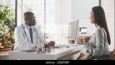 Bel giovane dottore africano sta avendo una conversazione con una paziente asiatica multietnica, parlando con lei durante la consultazione in una clinica sanitaria. Medico in cappotto bianco che lavora in ospedale moderno Foto Stock