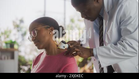 African Skin Care Professional utilizzando un Dermatoscopio per esaminare il tessuto del collo sulla pelle di una giovane donna nera attraente durante una visita di controllo salute in una clinica Foto Stock