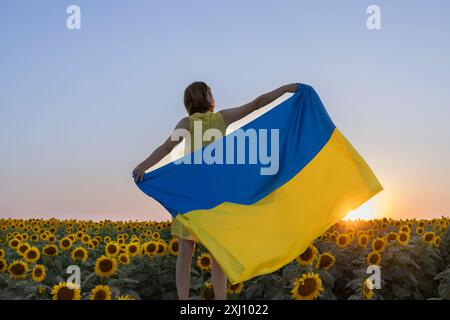 La donna tiene una grande bandiera Ucraina giallo-blu dietro di lei mentre si trova di fronte a un campo fiorito di girasoli. Siate orgogliosi di essere ucraini. Foto Stock