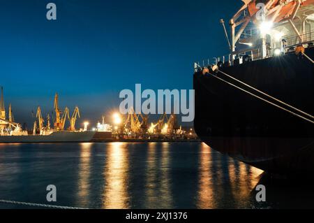 Notte al porto. Nave cargo con scafo nero ormeggiata al molo. Numerose gru portuali e navi sullo sfondo. Foto Stock