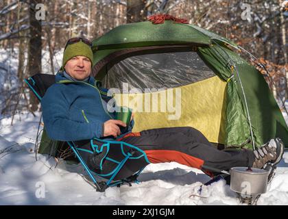 Un uomo vicino alla tenda si sta preparando a mangiare. Notte nella foresta invernale. per il sole, la foresta, la neve. Stile di vita attivo. Tenda nella neve. Inverno e viaggi Foto Stock