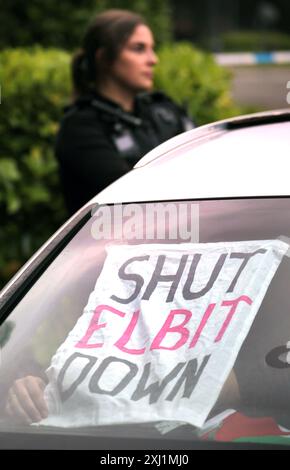 Bristol, Inghilterra, Regno Unito. 16 luglio 2024. Sul parabrezza è apposto un cartello con la scritta "Shut Elbit Down", mentre gli attivisti si attaccano alle serrature e bloccano la strada fuori dalla fabbrica di Elbit Systems con un'auto assistita da un agente di polizia. Per due giorni consecutivi gli attivisti hanno bloccato la strada che porta alla fabbrica di Elbit Systems impedendo ai lavoratori di frequentare i loro uffici. I sostenitori di Palestine Action usano i lock-on per rendere la loro rimozione più difficile e dispendiosa in termini di tempo ad Aztec West, Bristol. Essi sostengono che le armi prodotte da Elbit nel Regno Unito sono utilizzate dagli israeliani contro i pales Foto Stock