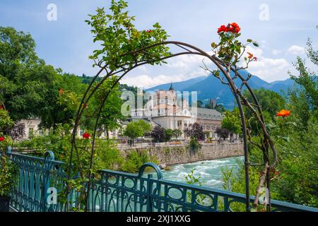 Merano, Südtirol, Italien - Kurhaus am Fluss Passer, an der Passerpromenade in der Altstadt. Vorne Rote Rosen auf der Thermenbrücke. Merano Süddtirol Italien *** Merano, alto Adige, Italy Spa House sul fiume Passer, sul lungomare di Passer nel centro storico Rose rosse di fronte al ponte termale Merano alto Adige Italia Foto Stock