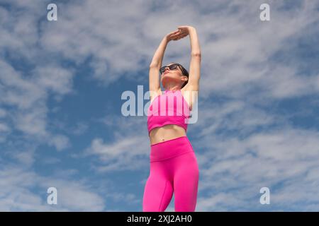 Donna sportiva e in forma che fa esercizi di stretching del braccio indossando abiti sportivi rosa contro il cielo blu Foto Stock