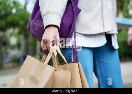 Primo piano di una donna che porta borse della spesa di carta marrone. Outfit elegante con borsetta viola, maglione bianco e jeans. Concetto di shopping sostenibile in urb Foto Stock