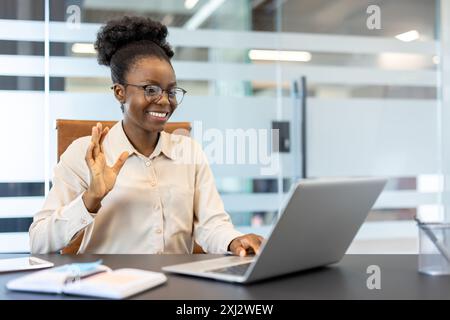 Donna d'affari sorridente che sta videochiamando con un laptop in ufficio. Donna professionista che saluta durante una riunione online. Concetto di lavoro remoto, tecnologia, comunicazione e connessioni aziendali. Foto Stock