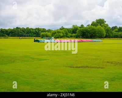 Colorate coperture su un vasto campo da cricket verde Roe Green Worsley sotto un cielo nuvoloso, circondato da alberi lussureggianti. Foto Stock