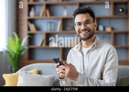 Un giovane sorridente che utilizza lo smartphone nel salotto moderno, seduto su un comodo divano. Luminoso e accogliente ambiente da ufficio, con libreria in legno e sfondo verde. Foto Stock