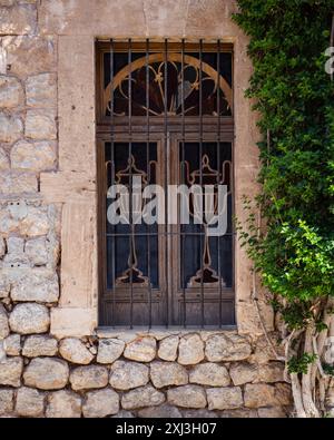 Una vecchia finestra in legno intagliato con barre in una parete di pietra chiara. Un cespuglio verde cresce nelle vicinanze. Mallorca, Spagna Foto Stock