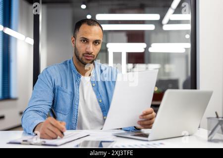 Ritratto di un serio ispanico che lavora in ufficio con laptop e documenti, annotando dati e informazioni. Guarda con sicurezza nella fotocamera. Foto Stock