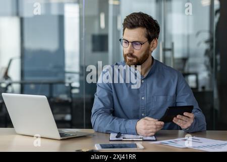 Contabile serio che si concentra sull'analisi finanziaria utilizzando notebook e tablet in un ufficio moderno. Focalizzata su documenti cartacei, dati e calcoli. Produttività concettuale, finanza e professionalità Foto Stock