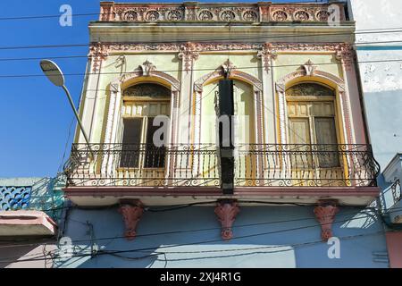 502 facciata di un edificio eclettico con primo piano dipinto di giallo pallido e modanature rosa, balcone con ringhiere in ferro su mensole scolpite floreali. Santiago-Cuba. Foto Stock