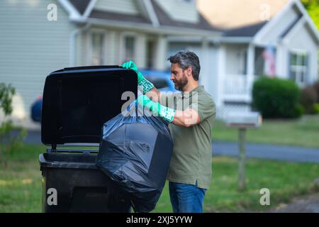 Un uomo spinge un grande cestino in plastica per pianificare lo smaltimento dei rifiuti settimanalmente. Uomo di mezza età che mette la spazzatura in un bidone della spazzatura in strada. Uomo Foto Stock