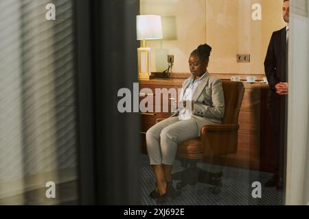Giovane direttrice finanziaria afroamericana o amministratore delegato in tuta formale seduta in poltrona in sala riunioni e messaggi di testo Foto Stock