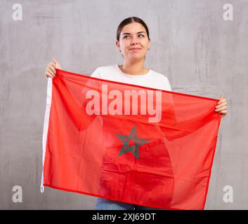 Giovane donna felice in posa e sorridente, in piedi con la bandiera del Marocco contro il muro grigio Foto Stock