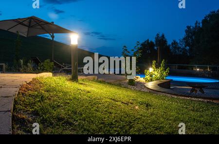 Vista serale di una lussuosa area piscina illuminata da luci LED contemporanee, con piscina, vialetto, ombrellone e vegetazione circostante, stile enfatizzato Foto Stock