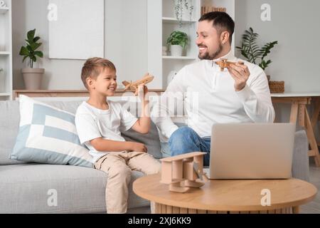 Figlio felice che tiene in mano aerei di legno e suo padre che lavora con un computer portatile sul divano a casa Foto Stock