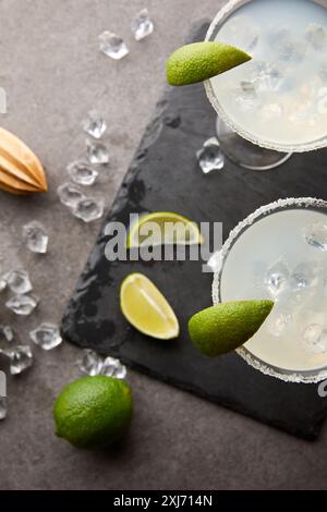 Vista dall'alto di alcol cocktail Margarita con pezzi di calce, cubetti di ghiaccio e spremitore in legno grigio sul piano portapaziente Foto Stock