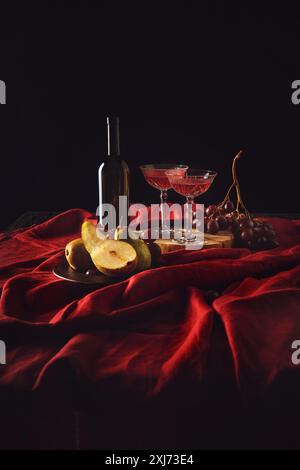 Natura morta con pere, uva e vino in bottiglia e bicchieri di cristallo su drappo rosso su nero Foto Stock