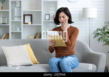Turbata e preoccupata giovane donna che legge una lettera ricevuta a casa, seduta sul divano e tenendo la testa. Foto Stock