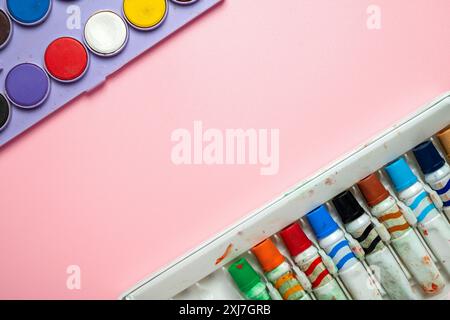 Una scatola di pastelli e una serie di acquerelli sono su sfondo rosa. I pastelli sono di vari colori e gli acquerelli sono in un vassoio Concetto di c Foto Stock