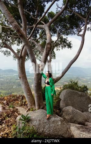 Una donna con un abito verde si appoggia su un grande albero sulla cima di una collina che si affaccia su un paesaggio verde lussureggiante. Foto Stock