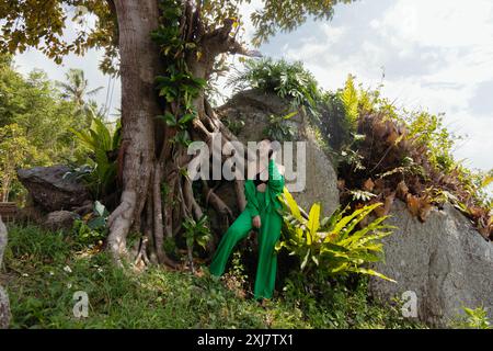 La donna posa accanto a un grande albero in un ambiente tropicale. Indossa un vestito verde ed è circondata da lussureggianti fogliame e rocce. Foto Stock