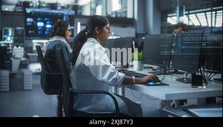 Diversi giovani colleghi indiani che lavorano su computer in un laboratorio di ricerca. Concentratevi sul collega donna sviluppatrice di software che lavora a una soluzione per il loro progetto di tecnologia industriale collaborativa Foto Stock