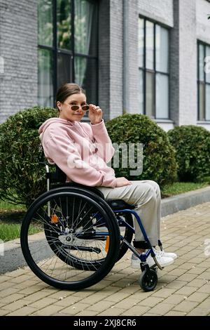 Una giovane donna con una felpa con cappuccio rosa siede su una sedia a rotelle all'aperto, sorridendo e regolando gli occhiali da sole. Foto Stock