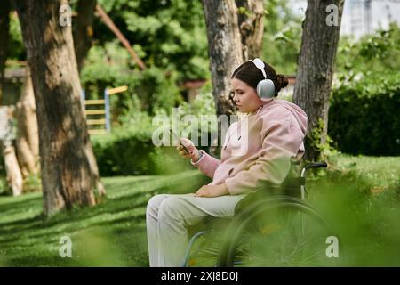 Una giovane donna in sedia a rotelle ascolta la musica sul suo telefono mentre è seduta in un parco. Indossa le cuffie e una felpa con cappuccio rosa. Foto Stock