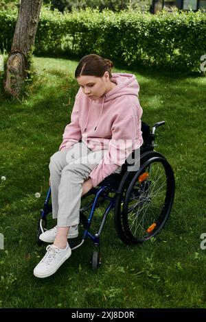 Una giovane donna siede su una sedia a rotelle in un parco erboso con una felpa con cappuccio rosa. Foto Stock