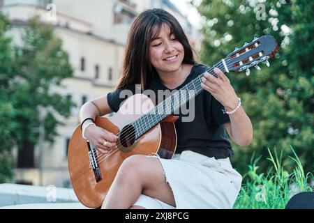 giovane latina che suona la chitarra in una città. donna seduta su una panchina di pietra che pratica con il suo strumento musicale. Foto Stock