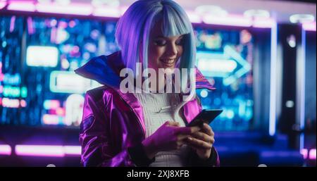 Ritratto di un'elegante Cyberpunk Cosplay Girl con capelli blu utilizzando lo smartphone. Giovane, felice, bella gamer Girl che naviga su Internet, chatta e fa shopping online in una futuristica camera al neon Foto Stock