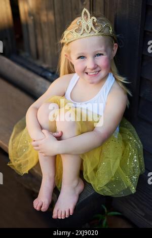 Sorridi, ritratto e bambino in abito da principessa per fantasia, giardino di casa e recinzione in legno. Ragazza felice in tutu, abiti da favola e corona di moda Foto Stock