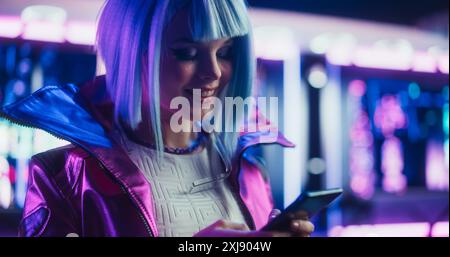 Ritratto di un'elegante Cyberpunk Cosplay Girl con capelli blu utilizzando lo smartphone. Giovane, felice, bella gamer Girl che naviga su Internet, chatta e fa shopping online in una futuristica camera al neon Foto Stock