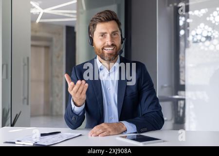 Un uomo d'affari allegro che indossa una cuffia interagisce durante una riunione aziendale virtuale, illustrando la sicurezza e l'accessibilità in un ambiente moderno. Foto Stock